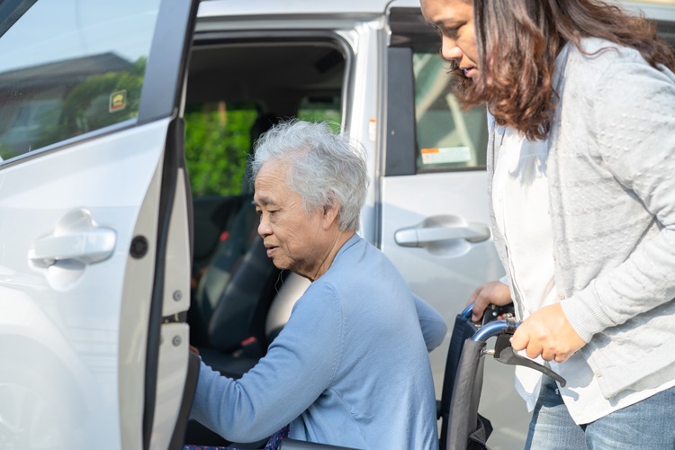 A care giver helps an elderly lady into a car