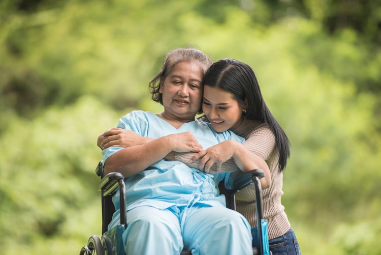 An elderly woman in a wheelchair being hugged by a young woman.