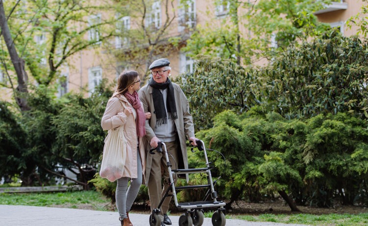 An elderly man walking with a young lady and using a walkin frame with wheels