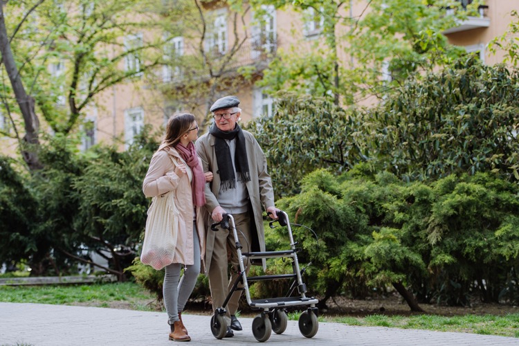 An elderly man walking with a young lady and using a walkin frame with wheels